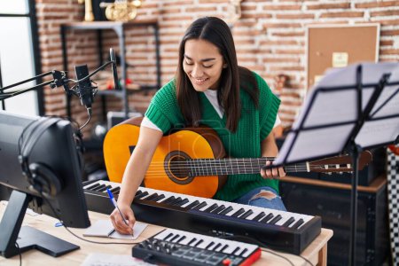 Photo for Young hispanic woman musician composing song playing classical guitar at music studio - Royalty Free Image
