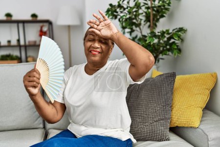 Foto de Mujer afroamericana mayor usando ventilador de mano sentado en el sofá en casa - Imagen libre de derechos