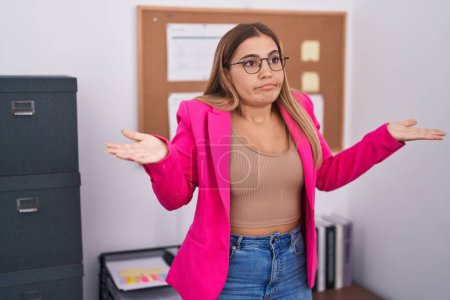 Photo for Young blonde woman standing at the office clueless and confused expression with arms and hands raised. doubt concept. - Royalty Free Image