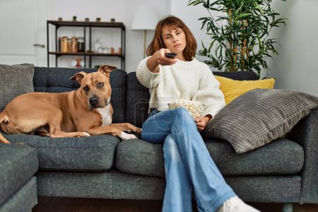 Téléchargez les photos : Jeune femme caucasienne regardant un film assis sur un canapé avec chien à la maison - en image libre de droit