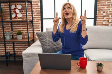 Photo for Middle age blonde woman using computer laptop at home amazed and surprised looking up and pointing with fingers and raised arms. - Royalty Free Image