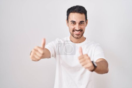 Téléchargez les photos : Bel homme hispanique debout sur fond blanc approuvant faire un geste positif avec la main, pouces levés souriant et heureux pour le succès. geste gagnant. - en image libre de droit