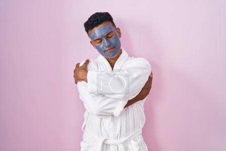 Téléchargez les photos : Jeune homme hispanique portant un masque de beauté et peignoir de bain se serrant dans ses bras heureux et positifs, souriant confiant. amour de soi et soins de soi - en image libre de droit