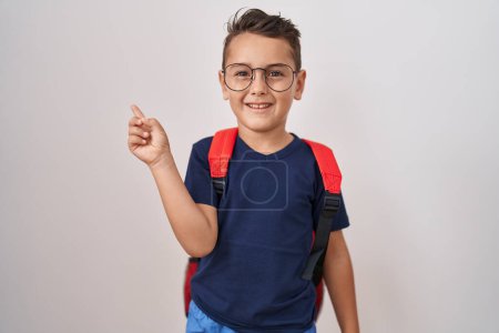 Foto de Pequeño niño hispano con gafas y mochila de estudiante sonriendo feliz señalando con la mano y el dedo hacia un lado - Imagen libre de derechos