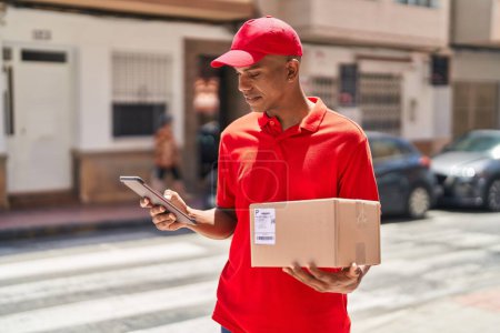 Photo for Young latin man delivery worker holding package using touchpad at street - Royalty Free Image