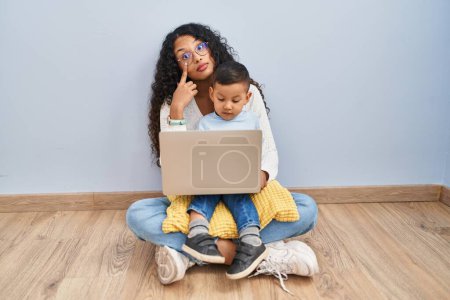 Téléchargez les photos : Jeune hispanique mère et enfant utilisant ordinateur portable assis sur le sol pointant vers l'oeil vous regarder geste, expression suspecte - en image libre de droit