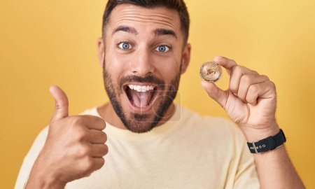 Foto de Hombre hispano guapo sosteniendo uniswap moneda criptomoneda sonriendo feliz y positivo, pulgar hacia arriba haciendo excelente y signo de aprobación - Imagen libre de derechos