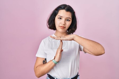 Photo for Young hispanic woman wearing casual white t shirt over pink background doing time out gesture with hands, frustrated and serious face - Royalty Free Image