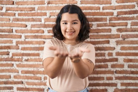 Foto de Mujer hispana joven de pie sobre la pared de ladrillos sonriendo con las palmas de las manos juntas recibiendo o dando gesto. retención y protección - Imagen libre de derechos