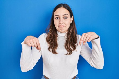 Photo for Young hispanic woman standing over blue background pointing down looking sad and upset, indicating direction with fingers, unhappy and depressed. - Royalty Free Image