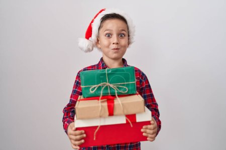 Téléchargez les photos : Petit garçon hispanique portant un chapeau de Noël tenant des cadeaux joues gonflées avec un visage drôle. bouche gonflée d'air, attrapant l'air. - en image libre de droit