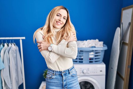 Foto de Joven mujer rubia sonriendo confiado abrazándose a sí mismo en la lavandería - Imagen libre de derechos