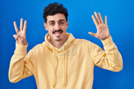 Photo for Hispanic man standing over blue background showing and pointing up with fingers number eight while smiling confident and happy. - Royalty Free Image