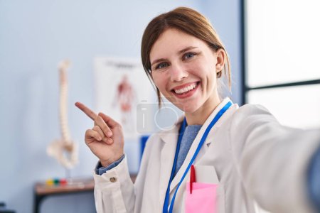Foto de Joven fisioterapeuta mujer que trabaja en la clínica de recuperación del dolor sonriendo feliz señalando con la mano y el dedo hacia un lado - Imagen libre de derechos