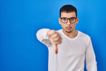 Téléchargez les photos : Jeune homme arabe portant une chemise blanche décontractée et des lunettes regardant malheureux et en colère montrant rejet et négatif avec les pouces vers le bas geste. mauvaise expression. - en image libre de droit