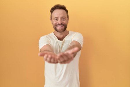 Photo for Middle age man with beard standing over yellow background smiling with hands palms together receiving or giving gesture. hold and protection - Royalty Free Image