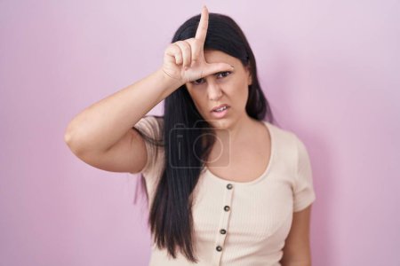 Photo for Young hispanic woman standing over pink background making fun of people with fingers on forehead doing loser gesture mocking and insulting. - Royalty Free Image