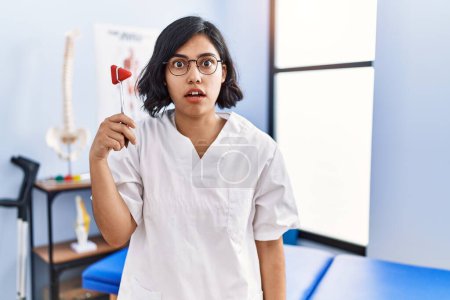Photo for Young hispanic physiotherapist woman holding reflex hammer scared and amazed with open mouth for surprise, disbelief face - Royalty Free Image
