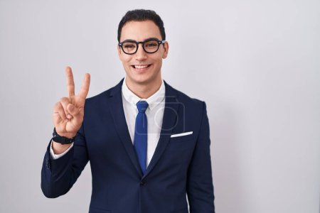Foto de Joven hombre hispano vistiendo traje y corbata mostrando y señalando con los dedos número dos mientras sonríe confiado y feliz. - Imagen libre de derechos