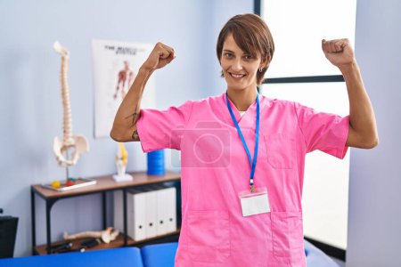 Téléchargez les photos : Femme brune travaillant à la clinique de réadaptation montrant les muscles des bras souriant fier. concept de fitness. - en image libre de droit
