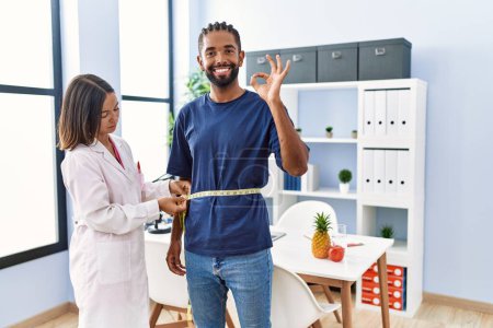 Photo for Dietitian at the clinic using tape measure measuring waist of client doing ok sign with fingers, smiling friendly gesturing excellent symbol - Royalty Free Image