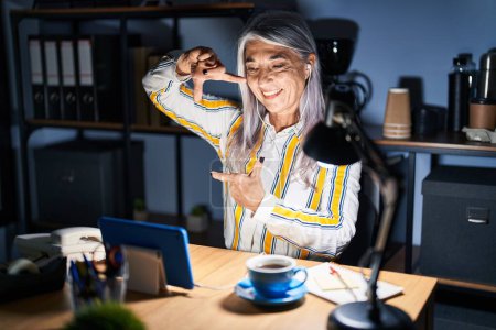 Téléchargez les photos : Femme d'âge moyen avec les cheveux gris travaillant au bureau la nuit souriant faisant cadre avec les mains et les doigts avec le visage heureux. concept de créativité et photographie. - en image libre de droit