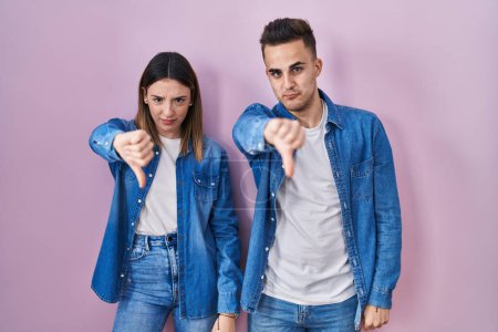 Photo for Young hispanic couple standing over pink background looking unhappy and angry showing rejection and negative with thumbs down gesture. bad expression. - Royalty Free Image