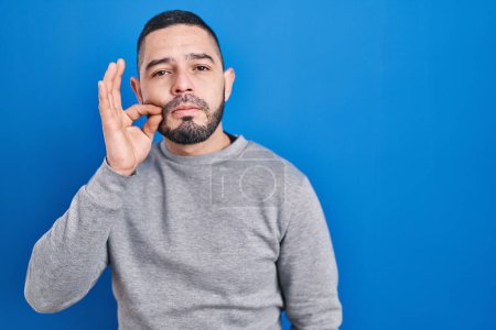 Téléchargez les photos : Homme hispanique debout sur fond bleu bouche et lèvres fermées comme zip avec les doigts. secret et silencieux, tabou parlant - en image libre de droit