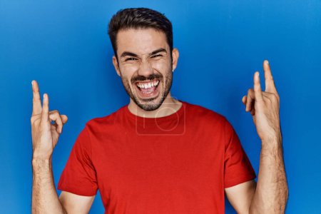 Foto de Joven hombre hispano con barba vistiendo camiseta roja sobre fondo azul gritando con expresión loca haciendo símbolo de roca con las manos arriba. Estrella de la música. concepto pesado. - Imagen libre de derechos