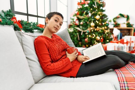 Foto de Joven mujer hispana leyendo libro y tomando café sentada junto al árbol de Navidad en casa - Imagen libre de derechos
