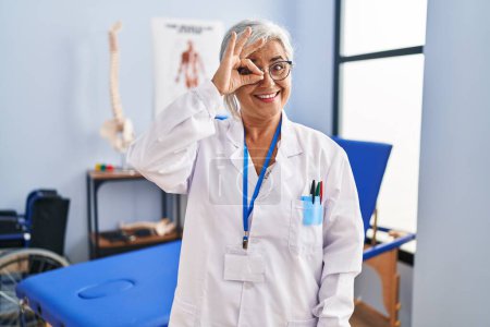Photo for Middle age woman with grey hair working at pain recovery clinic doing ok gesture with hand smiling, eye looking through fingers with happy face. - Royalty Free Image
