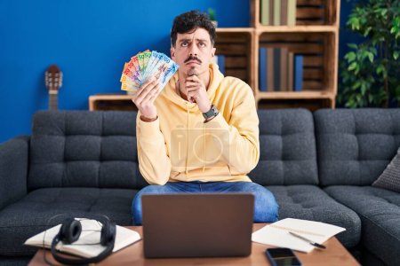 Photo for Hispanic man working with laptop holding swiss francs banknotes serious face thinking about question with hand on chin, thoughtful about confusing idea - Royalty Free Image