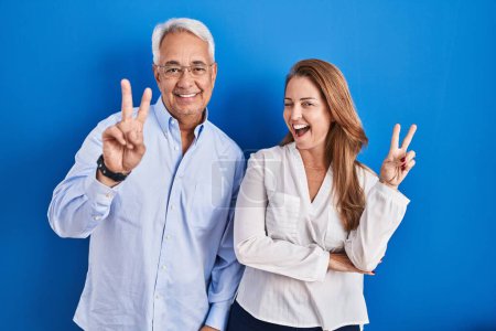 Téléchargez les photos : Couple hispanique du Moyen Age debout sur fond bleu souriant avec un sourire heureux clin d'oeil à la caméra faisant signe de victoire avec les doigts. numéro deux. - en image libre de droit