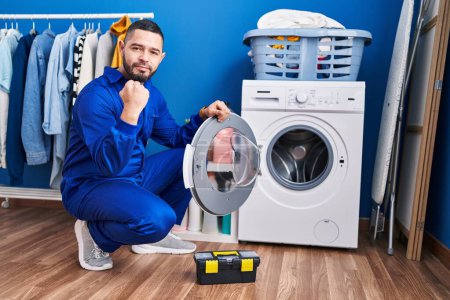 Téléchargez les photos : Réparateur hispanique travaillant sur la machine à laver criant fier, célébrant la victoire et le succès très excité avec les bras levés - en image libre de droit