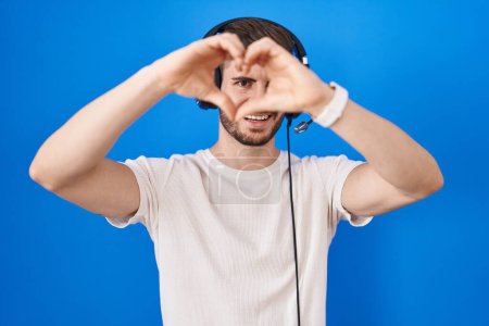 Téléchargez les photos : Homme hispanique avec barbe écoutant de la musique portant des écouteurs faisant forme de coeur avec la main et les doigts souriant en regardant à travers le signe - en image libre de droit