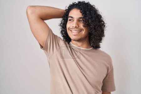 Téléchargez les photos : Homme hispanique avec les cheveux bouclés debout sur fond blanc souriant cheveux touchants confiants avec geste de la main vers le haut, posant attrayant et à la mode - en image libre de droit