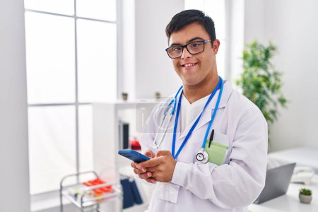 Téléchargez les photos : Homme trisomique portant l'uniforme de médecin en utilisant smartphone à la clinique - en image libre de droit