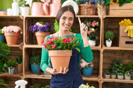 Foto de Joven mujer hispana trabajando en floristería sosteniendo planta sonriendo con una idea o pregunta señalando con el dedo con la cara feliz, número uno - Imagen libre de derechos