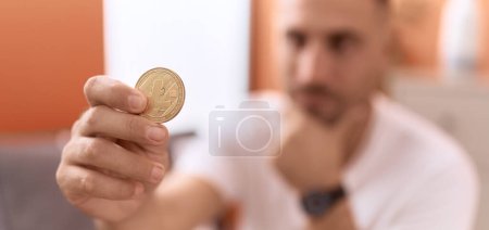 Foto de Joven hombre hispano sosteniendo moneda criptográfica litecoin sentado en el sofá en casa - Imagen libre de derechos