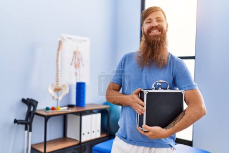 Téléchargez les photos : Jeune homme rousse portant un uniforme de physiothérapeute tenant une mallette à la clinique de physiothérapie - en image libre de droit