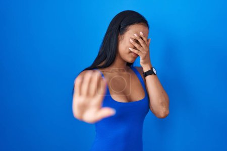 Téléchargez les photos : Femme hispanique debout sur fond bleu couvrant les yeux avec les mains et faisant arrêter geste avec expression triste et peur. concept embarrassé et négatif. - en image libre de droit