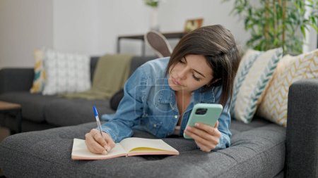 Téléchargez les photos : Jeune femme caucasienne utilisant l'écriture smartphone sur ordinateur portable à la maison - en image libre de droit