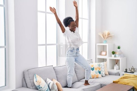 Téléchargez les photos : Femme afro-américaine souriante danse confiante sur le canapé à la maison - en image libre de droit
