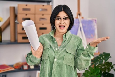 Téléchargez les photos : Jeune femme asiatique tenant du papier au studio d'art célébrant la réalisation avec sourire heureux et expression gagnante avec la main levée - en image libre de droit