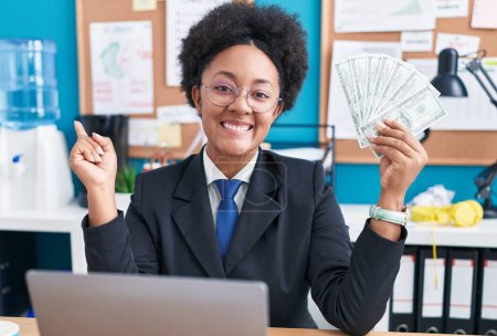 Foto de Hermosa mujer africana con el pelo rizado sosteniendo el dinero en la oficina sonriendo feliz señalando con la mano y el dedo a un lado - Imagen libre de derechos