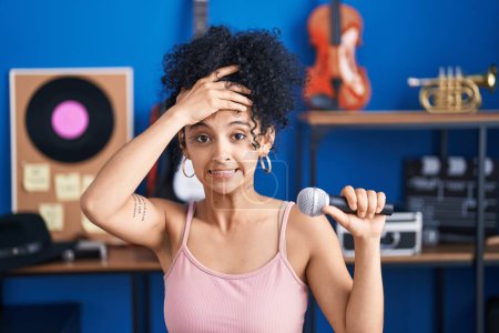 Téléchargez les photos : Femme hispanique avec des cheveux bouclés chantant chanson en utilisant le microphone au studio de musique stressé et frustré avec la main sur la tête, visage surpris et en colère - en image libre de droit