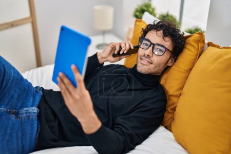 Foto de Hombre hispano joven usando touchpad hablando en el teléfono inteligente en el dormitorio - Imagen libre de derechos
