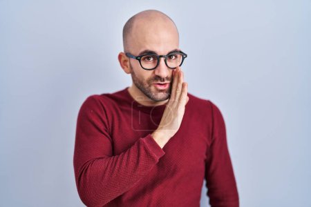 Photo for Young bald man with beard standing over white background wearing glasses hand on mouth telling secret rumor, whispering malicious talk conversation - Royalty Free Image