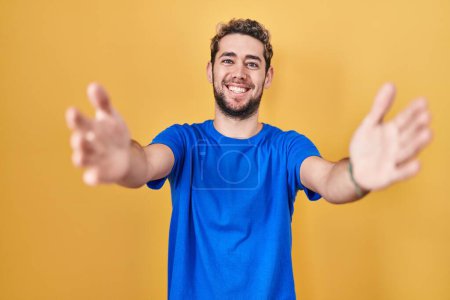 Téléchargez les photos : Hispanic man with beard standing over yellow background looking at the camera smiling with open arms for hug. cheerful expression embracing happiness. - en image libre de droit