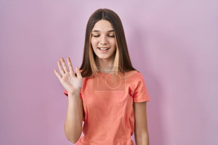 Foto de Adolescente chica de pie sobre rosa fondo renunciando a decir hola feliz y sonriente, gesto de bienvenida amistoso - Imagen libre de derechos